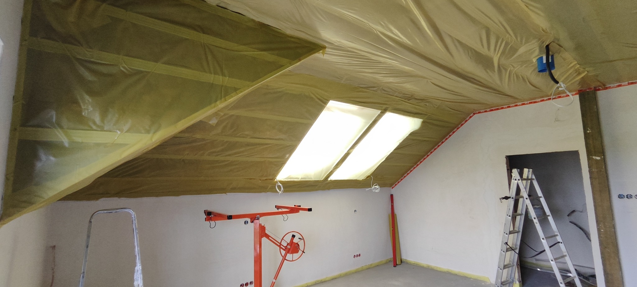 Poddasze w trakcie ocieplania, widoczne wełna mineralna zabezpieczona folią, okna dachowe, podnośnik do płyt gipsowych i drabina.