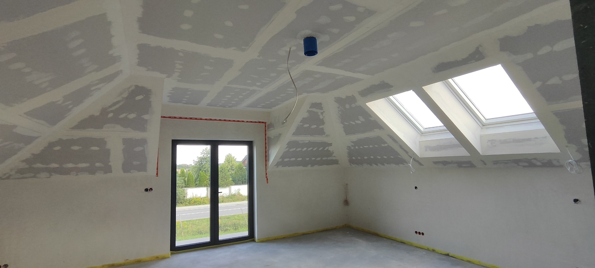Poddasze w trakcie wykańczania: widoczne płyty kartonowo-gipsowe na skośnym suficie i ścianach, szpachlowane łączenia, okna dachowe wpuszczające światło, przygotowane punkty elektryczne. Taśma...