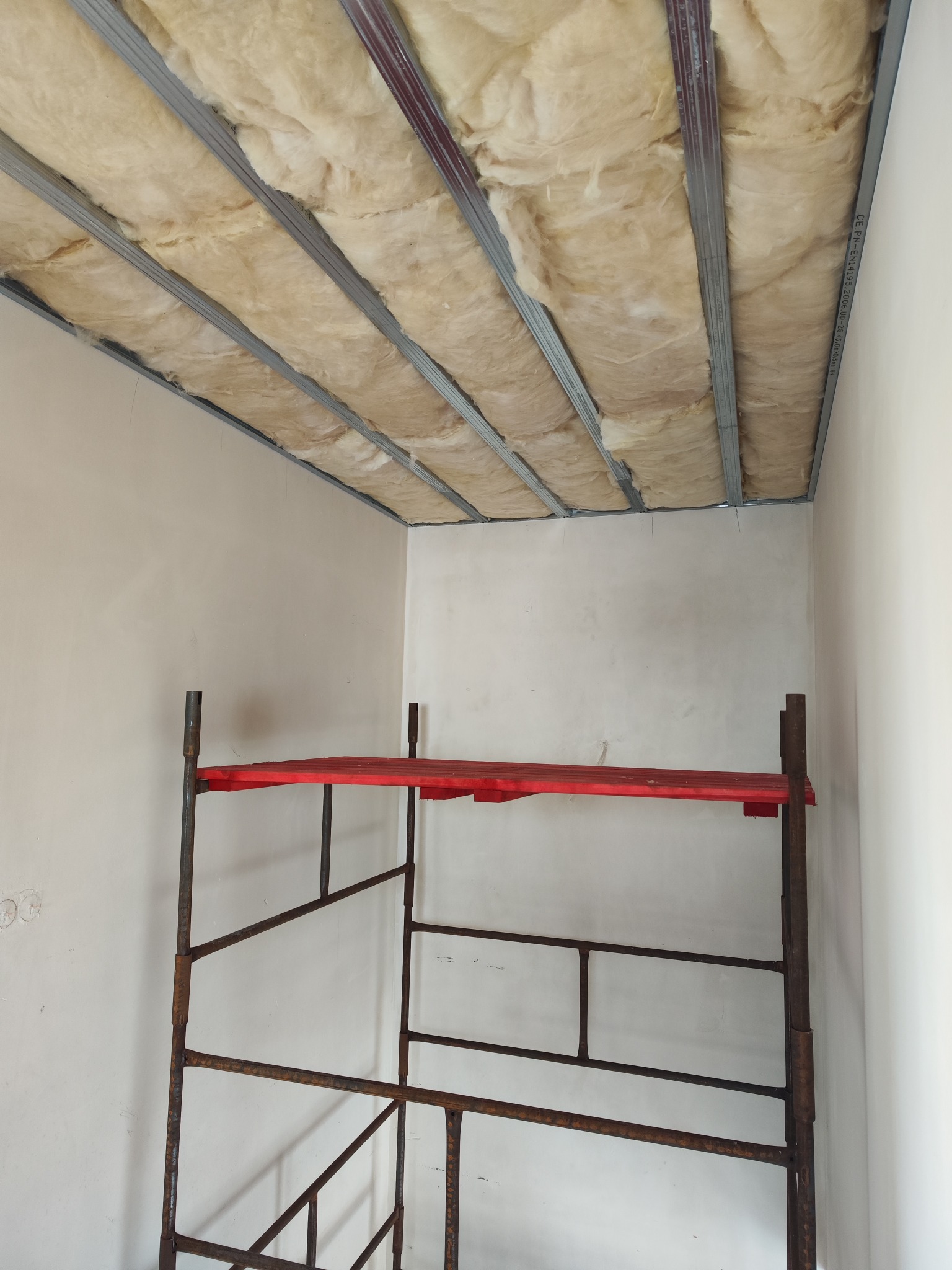 Szkielet metalowej konstrukcji sufitu z wypełnieniem z wełny mineralnej, widok z dołu, w tle fragment rusztowania malarskiego z czerwoną platformą.