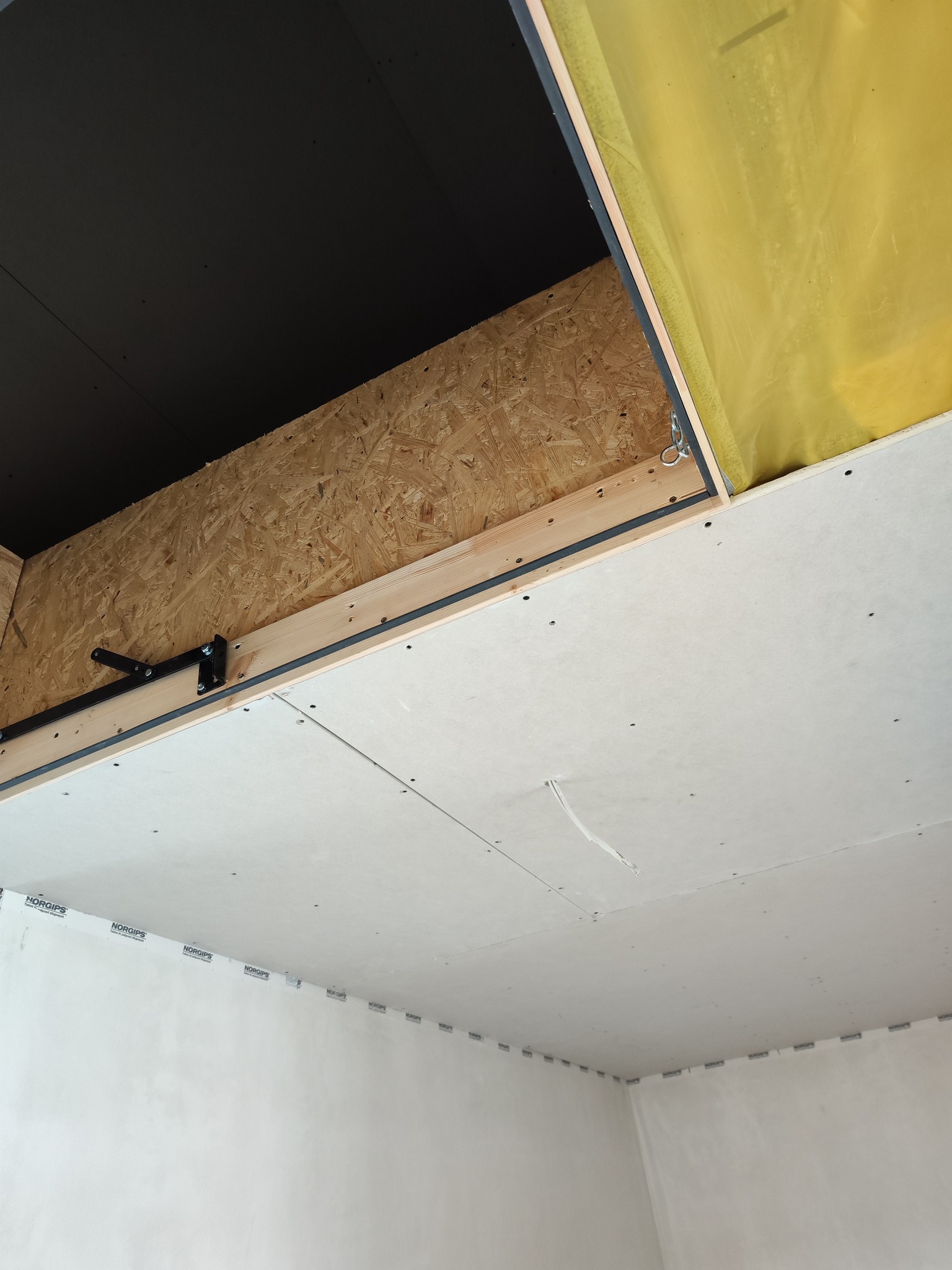 Montaż płyty gipsowo-kartonowej na suficie z widoczną izolacją z żółtej wełny mineralnej i elementami konstrukcyjnymi z drewna OSB, widok od dołu.