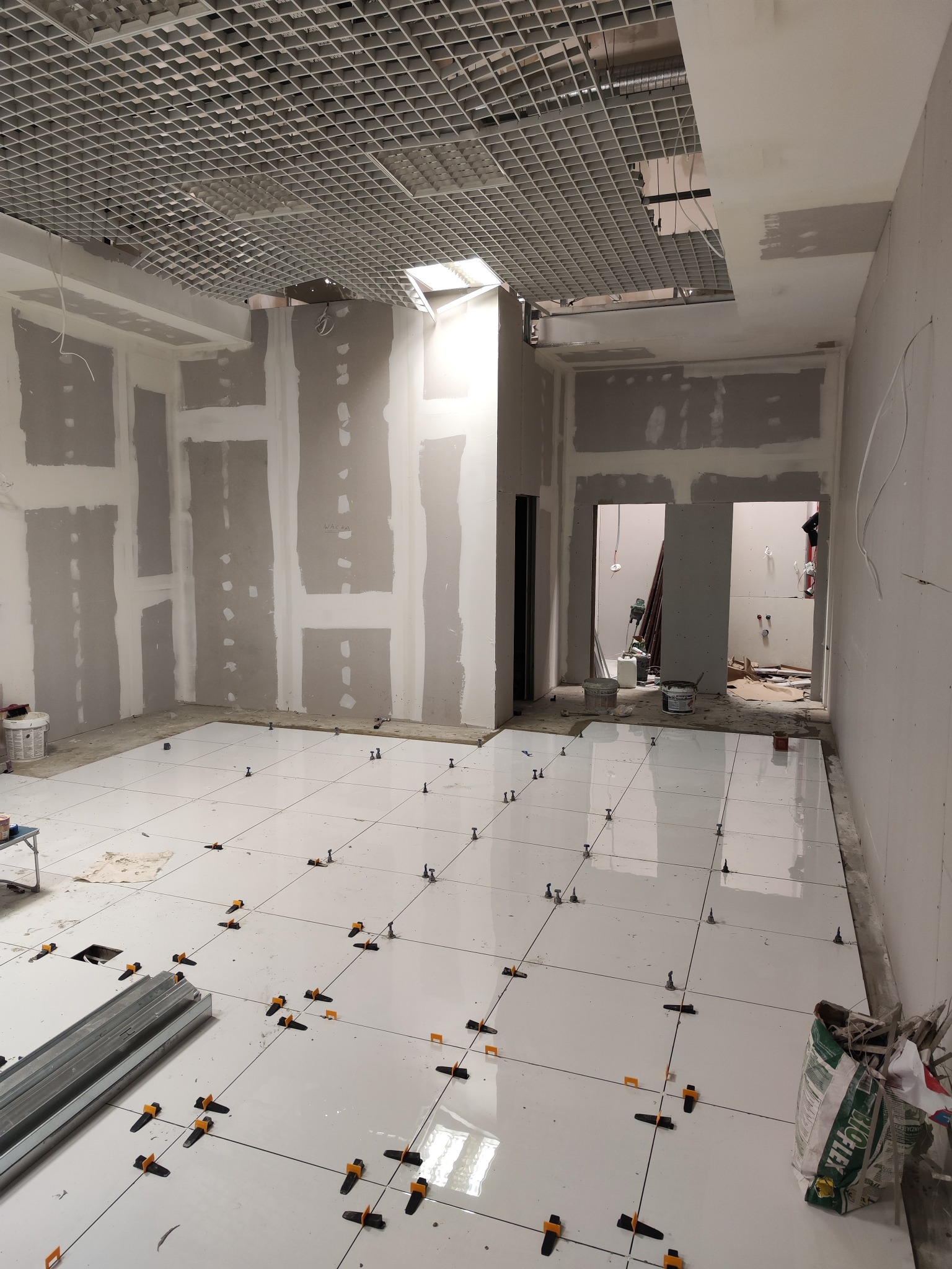 Wnętrze w trakcie remontu: białe płytki podłogowe z systemem poziomowania, ściany przygotowane pod gładzie, sufit z odsłoniętą konstrukcją metalową, widoczne materiały budowlane i narzędzia.