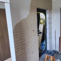 Wnętrze w trakcie prac wykończeniowych, fragment ściany z imitacją cegły, częściowo otynkowany, z widocznym przejściem do innego pomieszczenia zabezpieczonego folią, narzędzia i taboret na podłodze.