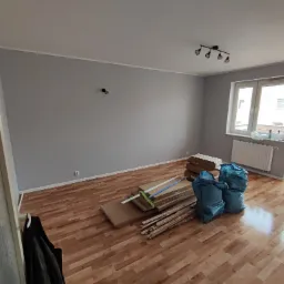 Pokój z parkietem w trakcie remontu, widoczne paczki z panelami, listwy przypodłogowe i worki na śmieci, ściany pomalowane na szaro, okno z widokiem na inne budynki.