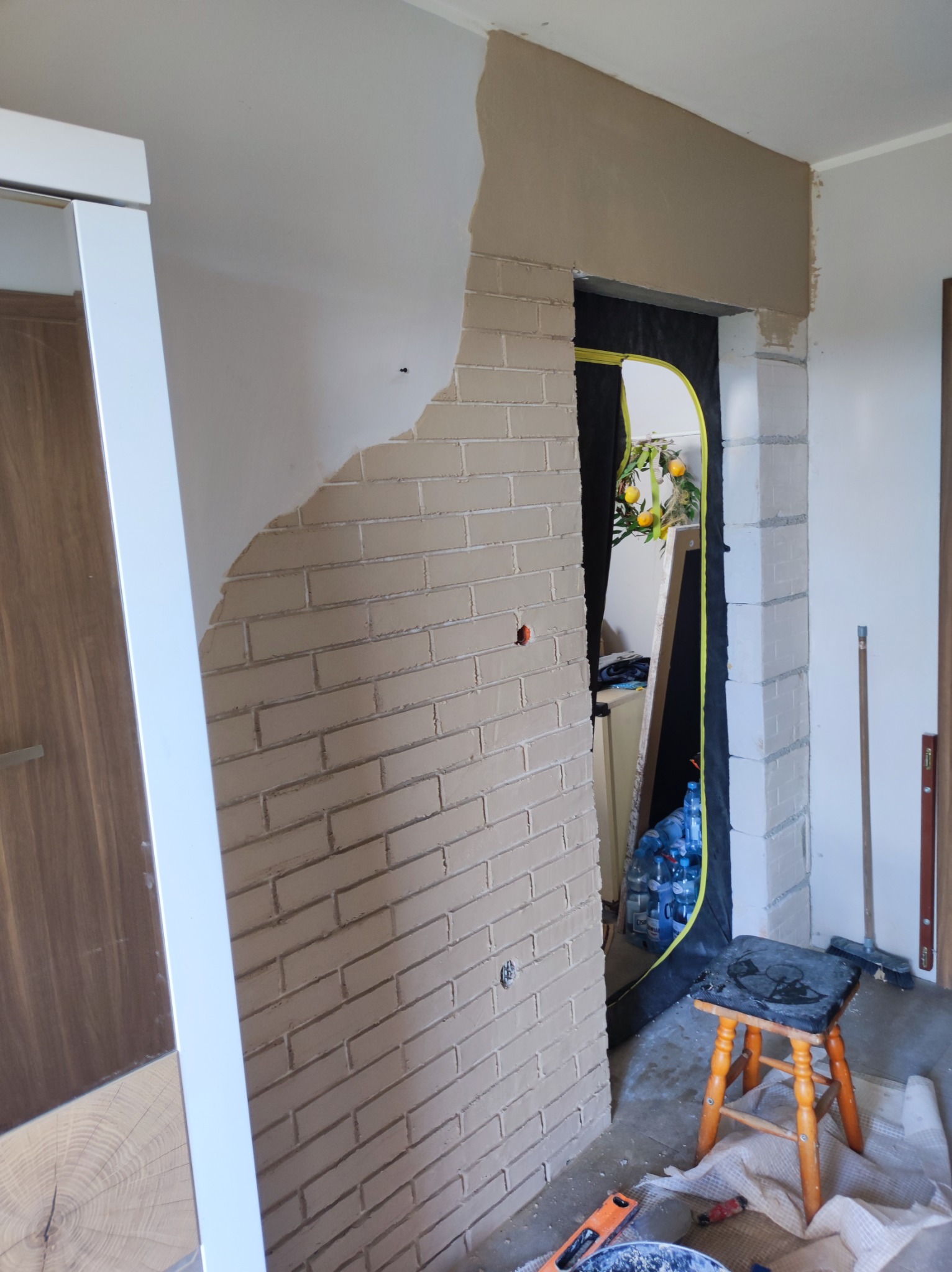 Wnętrze w trakcie prac wykończeniowych, fragment ściany z imitacją cegły, częściowo otynkowany, z widocznym przejściem do innego pomieszczenia zabezpieczonego folią, narzędzia i taboret na podłodze.