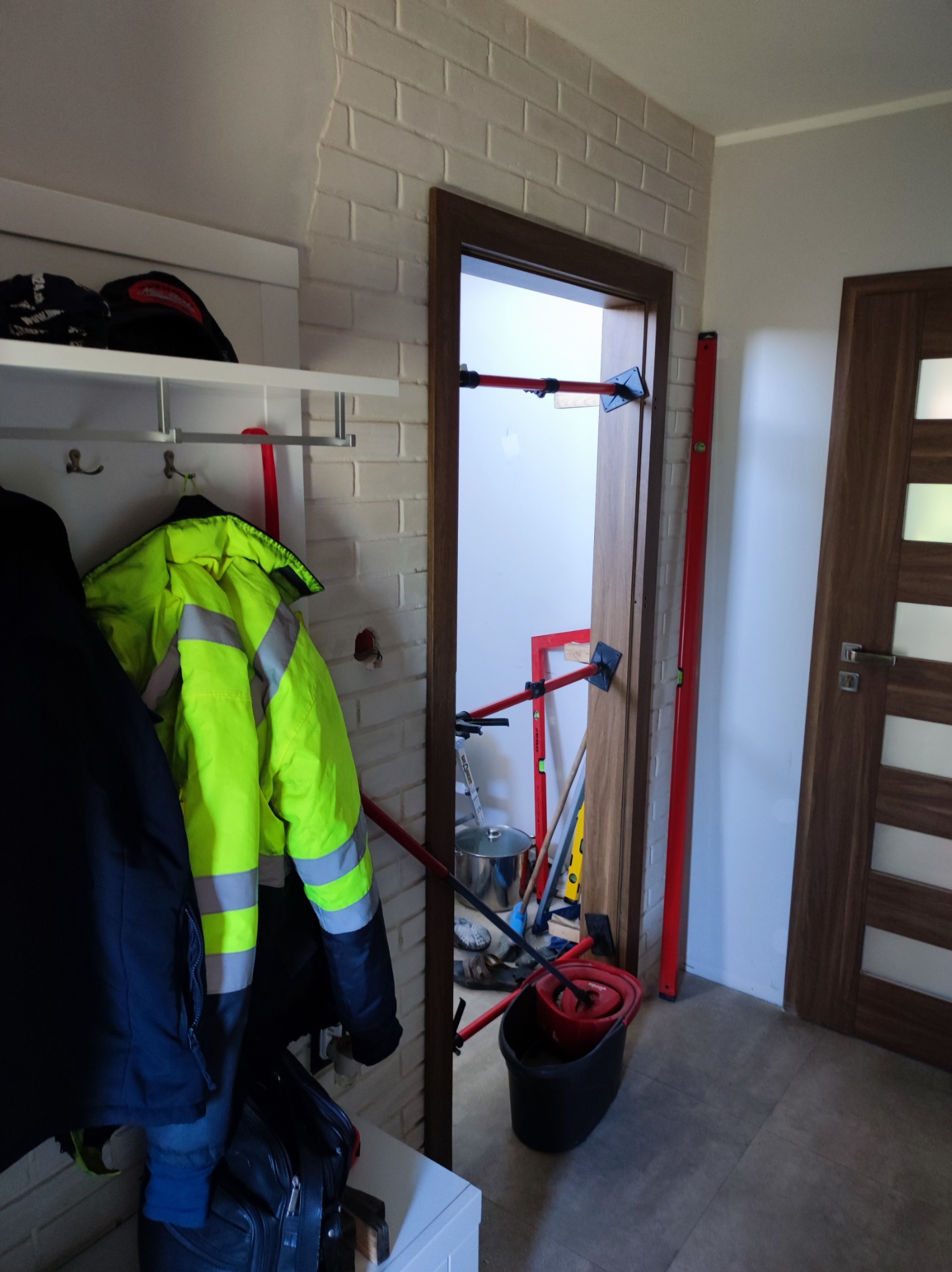 Wnętrze mieszkania w trakcie remontu: widoczny montaż ościeżnicy drzwiowej z użyciem rozpórek teleskopowych, poziomica oparta o ścianę, wiadro z mopem i narzędzia w tle.