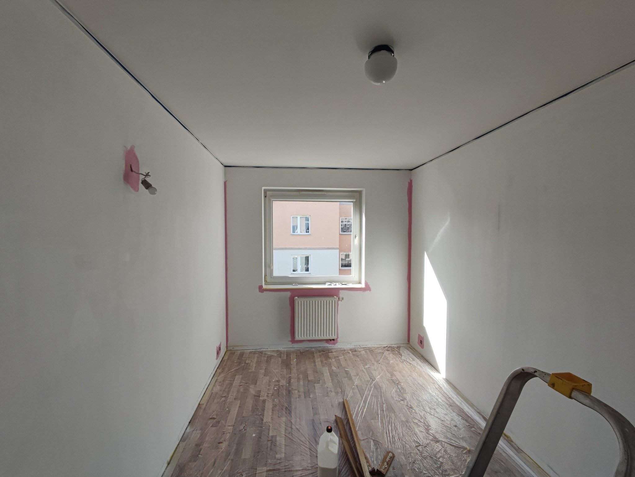 Pokój w trakcie przygotowania do malowania: świeżo pomalowane ściany na biało, zabezpieczone taśmą malarską krawędzie, podłoga przykryta folią ochronną, widoczna drabina i akcesoria malarskie.