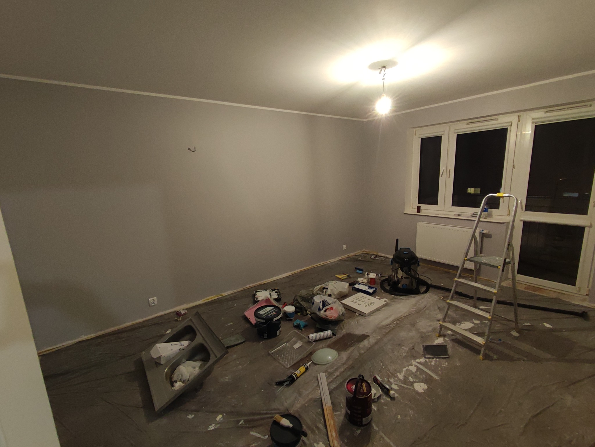Pokój po świeżym pomalowaniu ścian na szaro, zabezpieczona folią podłoga z rozłożonymi narzędziami malarskimi i puszkami farb, aluminiowa drabina oparta o okno.