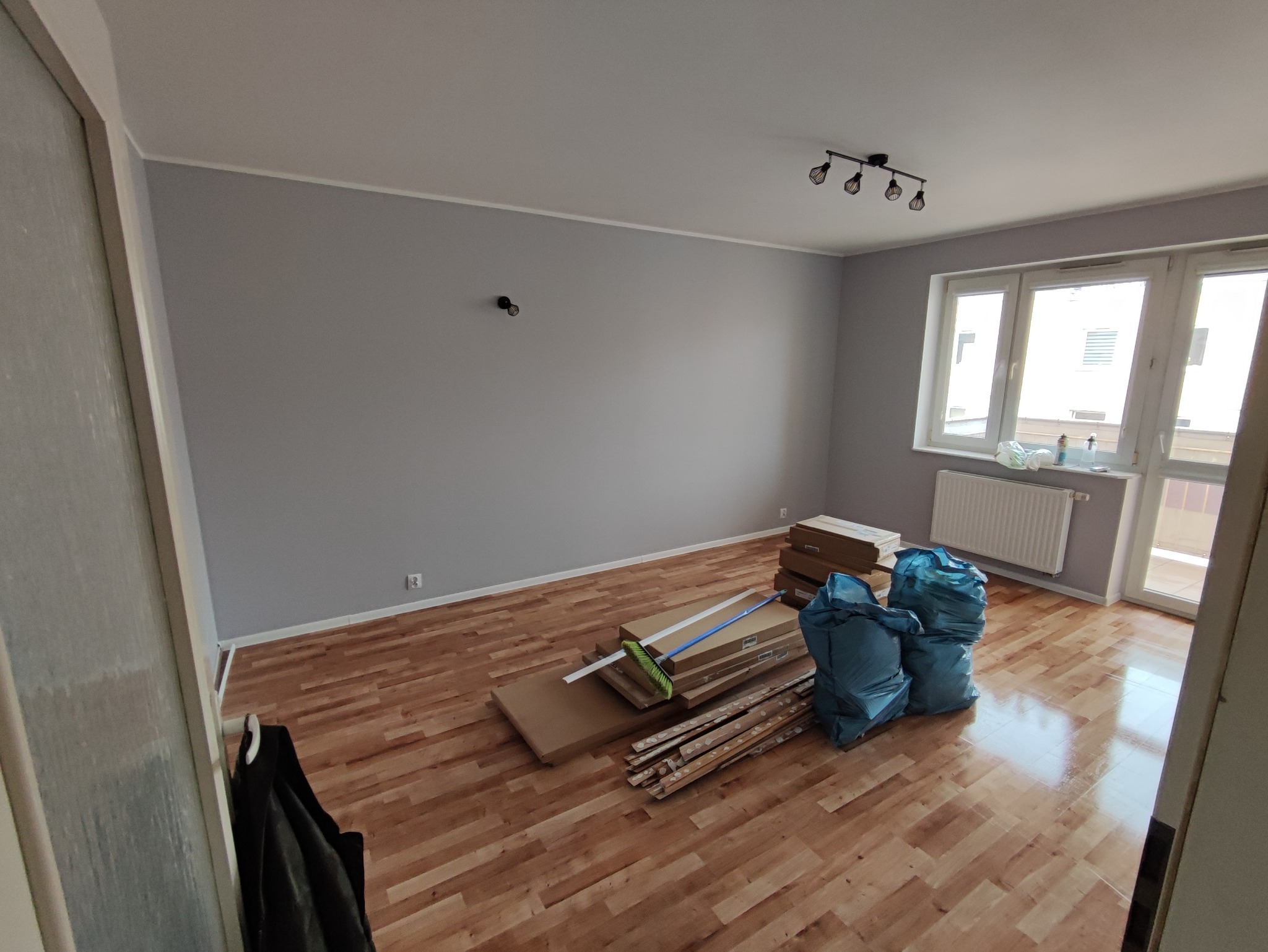 Pokój z parkietem w trakcie remontu, widoczne paczki z panelami, listwy przypodłogowe i worki na śmieci, ściany pomalowane na szaro, okno z widokiem na inne budynki.
