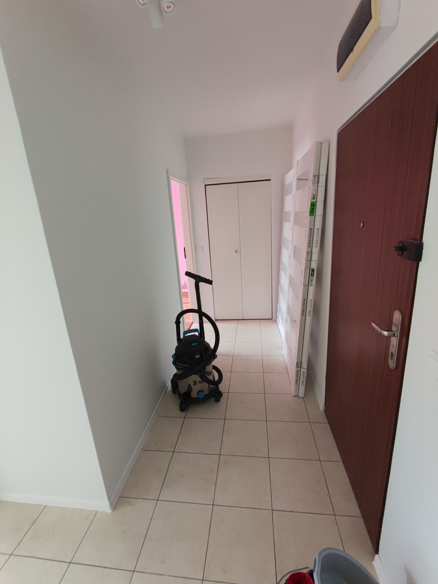 Korytarz w trakcie prac wykończeniowych, widoczny odkurzacz przemysłowy, zapakowane ościeżnice drzwiowe Stira-Door oparte o ścianę, oraz brązowe drzwi wejściowe.