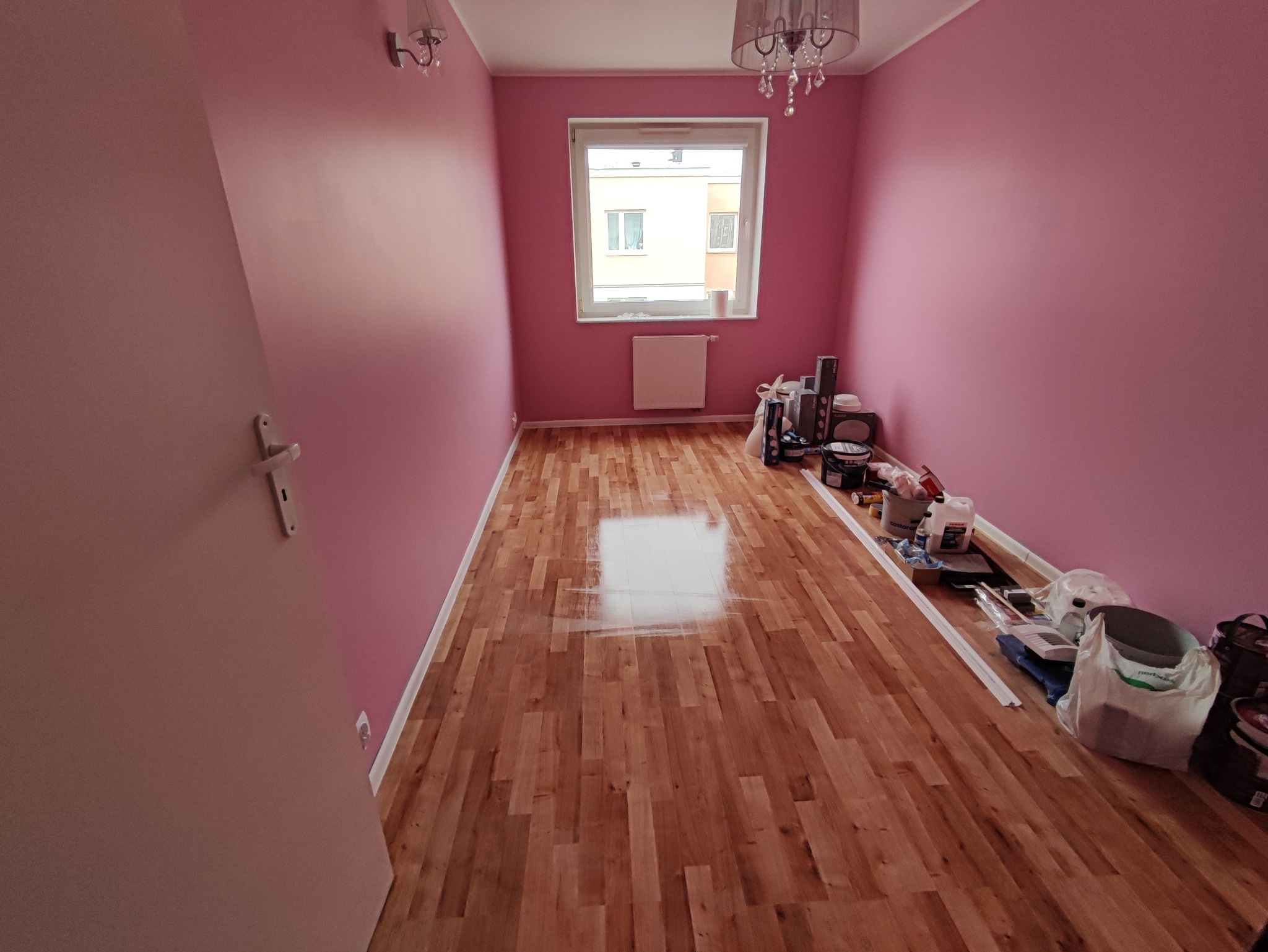 Pokój po malowaniu na różowo, z drewnianą podłogą i widocznymi narzędziami malarskimi oraz puszkami z farbą w rogu pomieszczenia.
