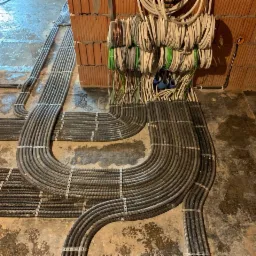 Szereg ułożonych równolegle, szarych rur karbowanych przytwierdzonych metalowymi uchwytami do podłoża i ściany z cegieł, z wystającymi przewodami elektrycznymi w tle, na surowym etapie budowy.