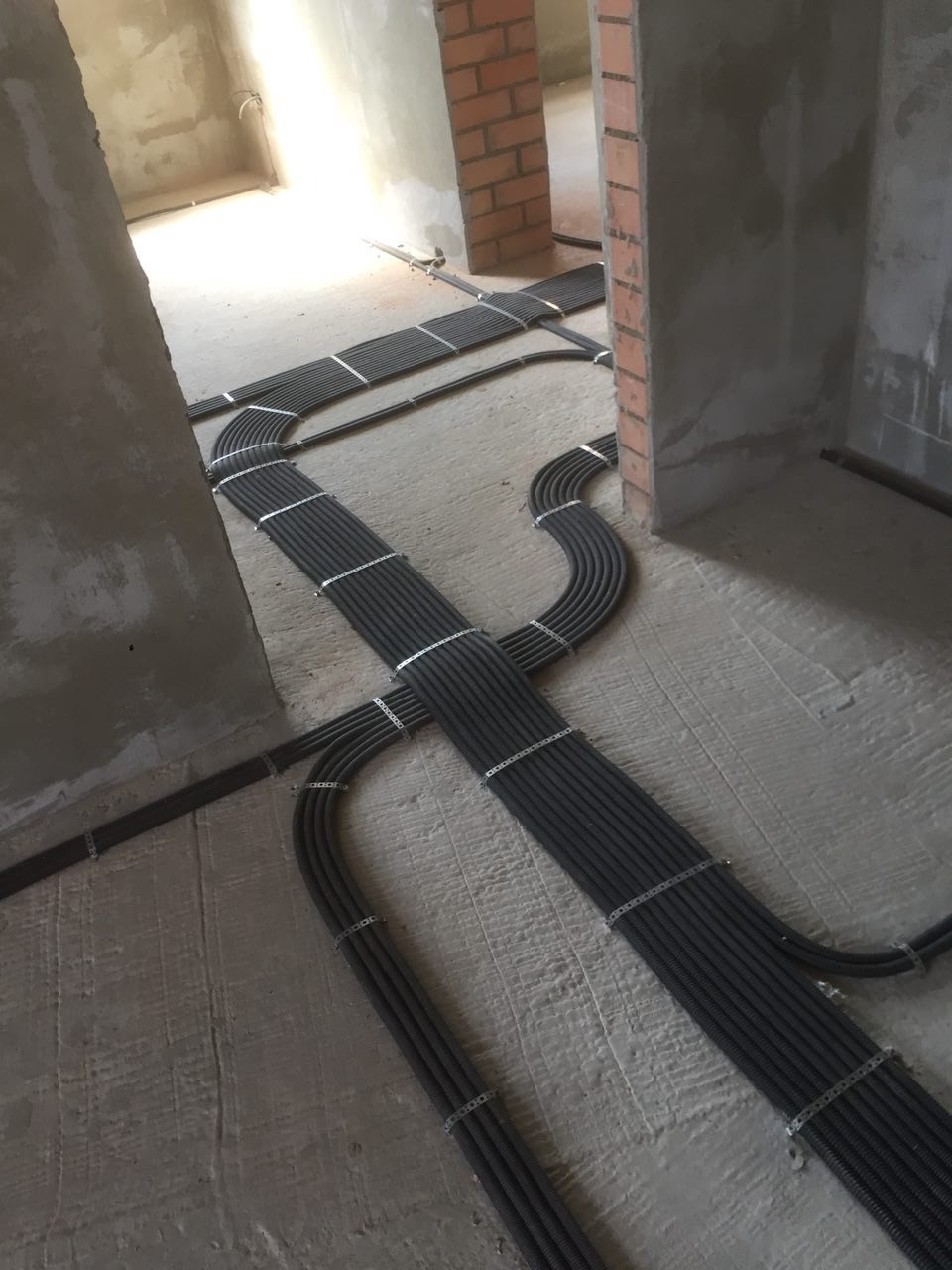 Rury instalacyjne ułożone na surowym betonowym podłożu w trakcie budowy, zabezpieczone metalowymi uchwytami, prowadzone wzdłuż ścian z surowej cegły.