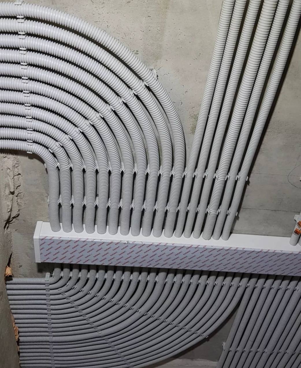 Równoległe rzędy szarych rurek karbowanych, ułożonych w łuk i prosto, przymocowanych do surowego betonowego sufitu i ściany za pomocą białej listwy montażowej.