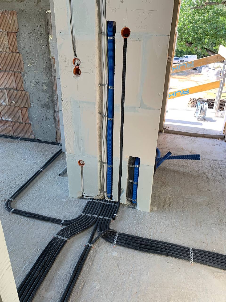 Instalacja elektryczna w trakcie montażu: liczne rury karbowane prowadzone w podłodze i ścianie, zabezpieczone metalowymi uchwytami, widoczne puszki instalacyjne w ścianie z gipsu.