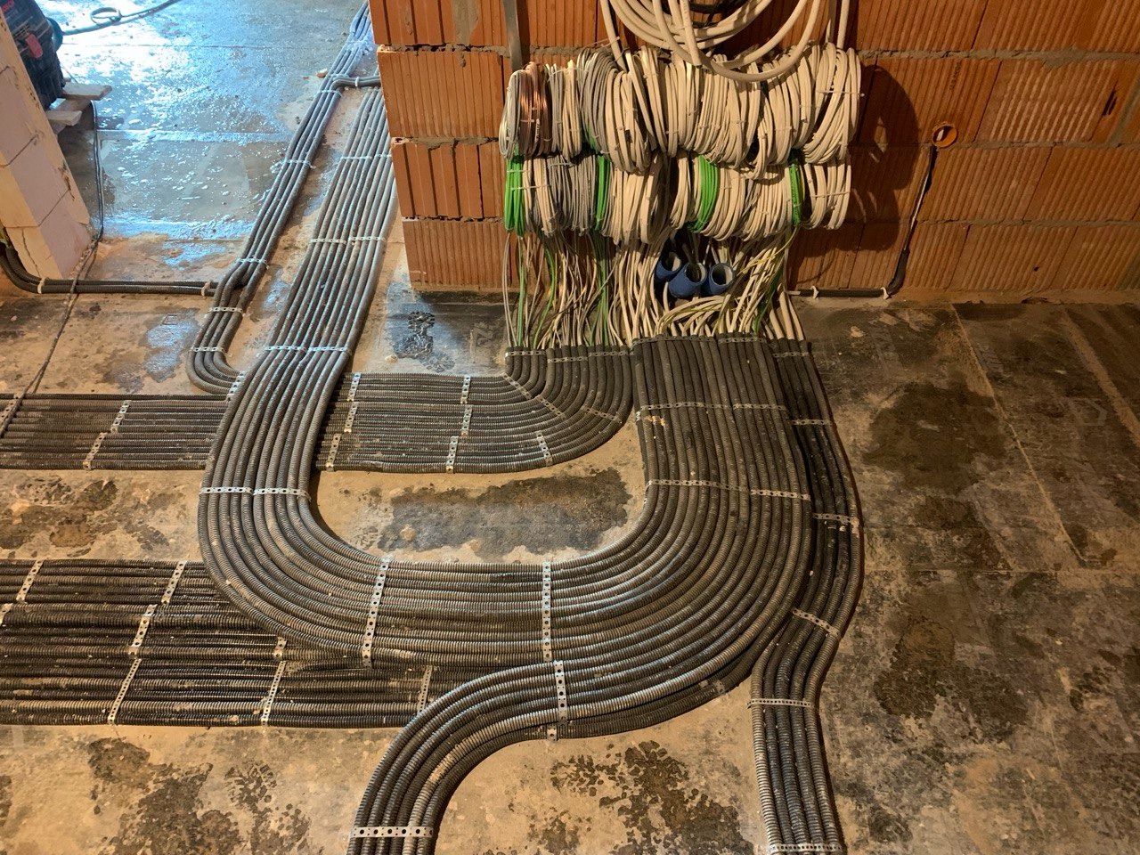 Szereg ułożonych równolegle, szarych rur karbowanych przytwierdzonych metalowymi uchwytami do podłoża i ściany z cegieł, z wystającymi przewodami elektrycznymi w tle, na surowym etapie budowy.