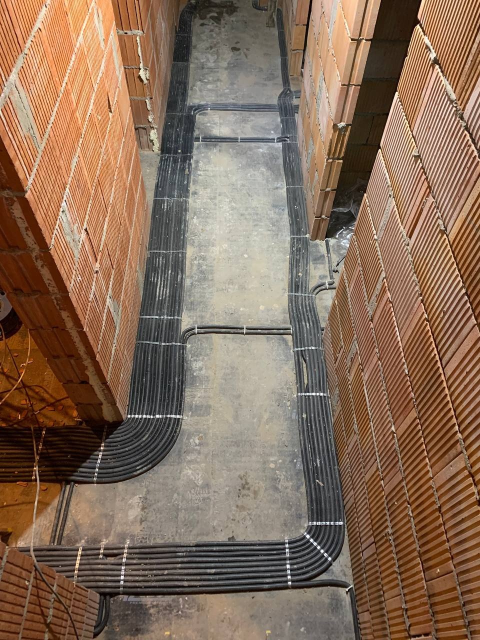 Równolegle ułożone rury instalacyjne, zabezpieczone białymi uchwytami, prowadzone wzdłuż ceglanych ścian w surowym stanie.