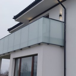 IronPeak Jarosław Sawic - Nowoczesny balkon z matową szklaną osłoną i czarną ramą, ozdobiony delikatnymi lampkami, przymocowany do białej ściany budynku z czarnym dachem.