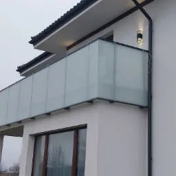 Nowoczesny balkon z matową szklaną osłoną i czarną ramą, ozdobiony delikatnymi lampkami, przymocowany do białej ściany budynku z czarnym dachem.