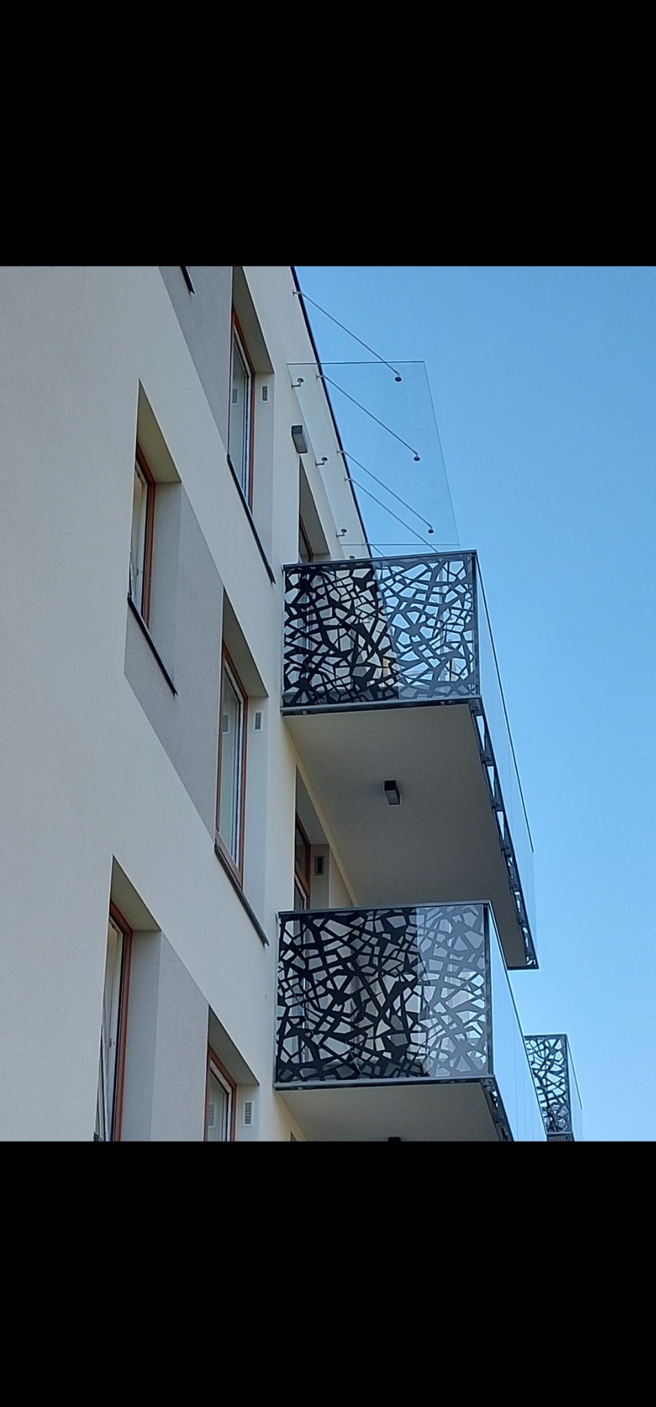 Nowoczesne balkony z ażurowymi balustradami i szklanymi zadaszeniami, widok z dołu na tle błękitnego nieba.