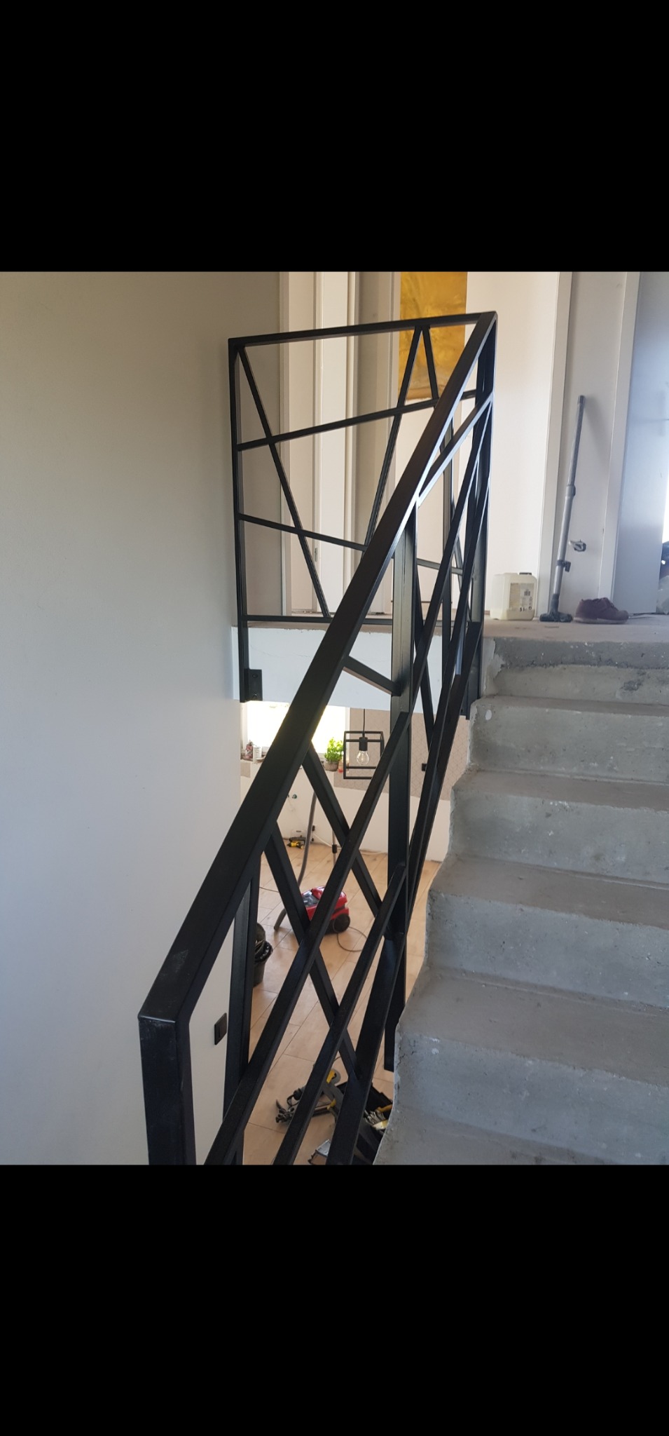 Nowoczesna, czarna balustrada schodowa o geometrycznym wzorze, zamontowana na betonowych schodach w surowym wnętrzu. Widoczne narzędzia i sprzęt budowlany sugerują trwający remont.