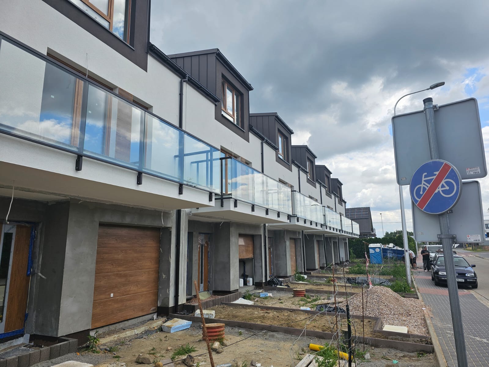 Rząd nowych domów szeregowych z balkonami zabezpieczonymi szklanymi barierkami, w trakcie wykończenia terenu wokół budynków.