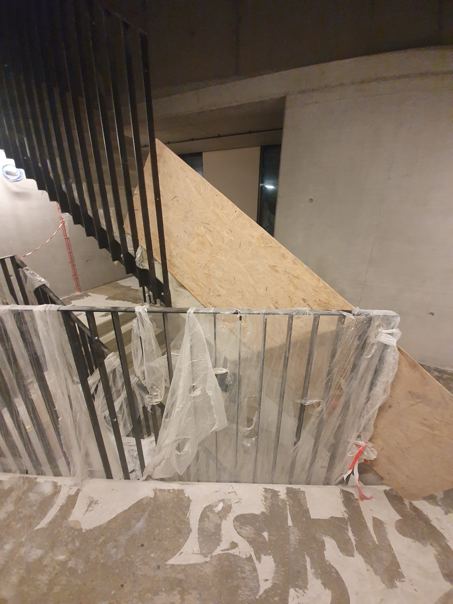 Częściowo zamontowana balustrada schodowa z czarnych, pionowych prętów i poziomych poręczy, zabezpieczona folią ochronną w trakcie prac wykończeniowych na betonowych schodach.