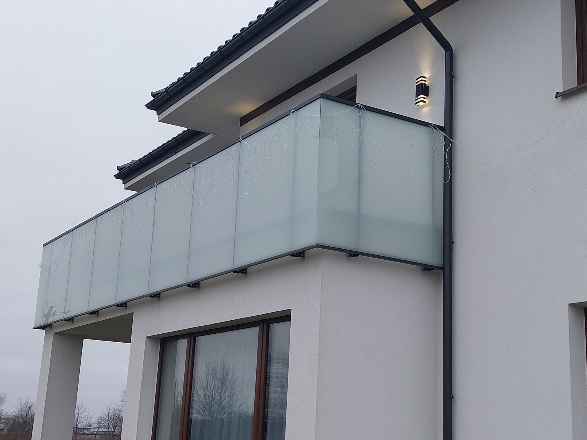 Nowoczesny balkon z matową szklaną osłoną i czarną ramą, ozdobiony delikatnymi lampkami, przymocowany do białej ściany budynku z czarnym dachem.