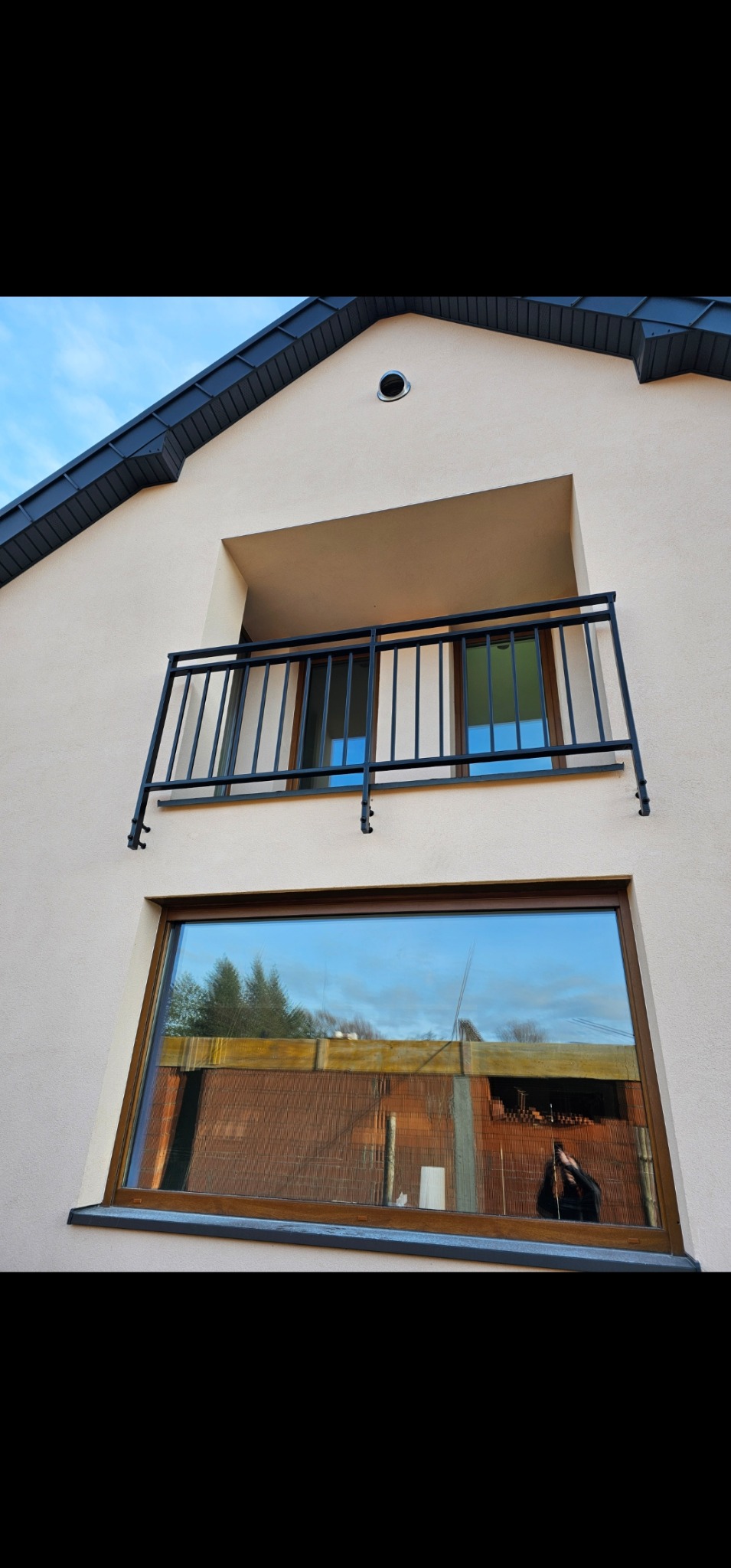 Czarna, metalowa barierka balkonowa zamontowana na elewacji domu z widocznym oknem, w którym odbija się budowa.
