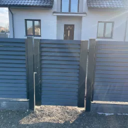 Nowoczesne ogrodzenie panelowe z poziomych, szarych paneli, zamontowane na betonowej podmurówce, z widocznym domem jednorodzinnym w tle, oświetlone jasnym światłem słonecznym.