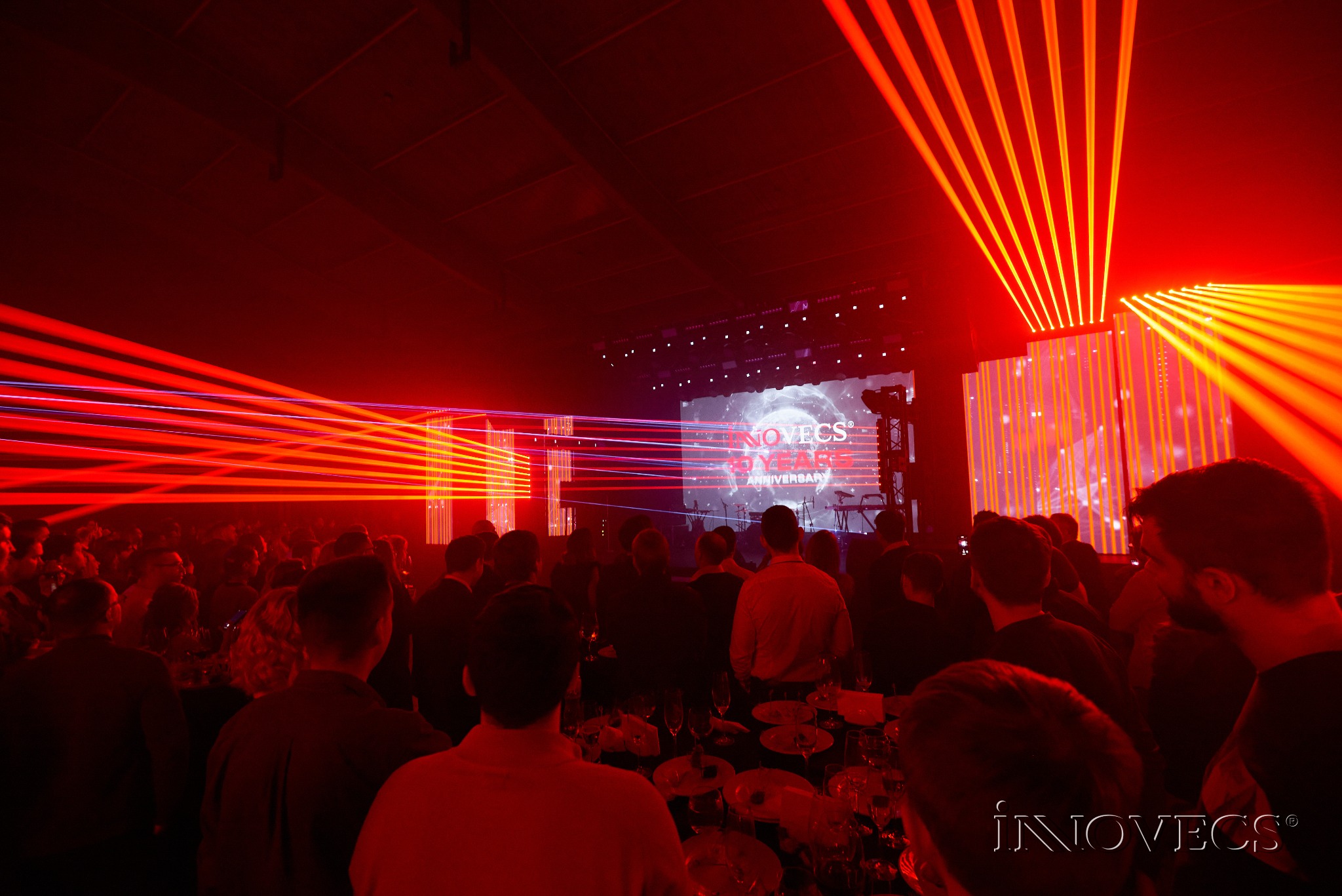 Widok z tyłu na tłum ludzi podczas wydarzenia w hali z czerwonym oświetleniem laserowym, ekranem wyświetlającym logo i sceną w tle.