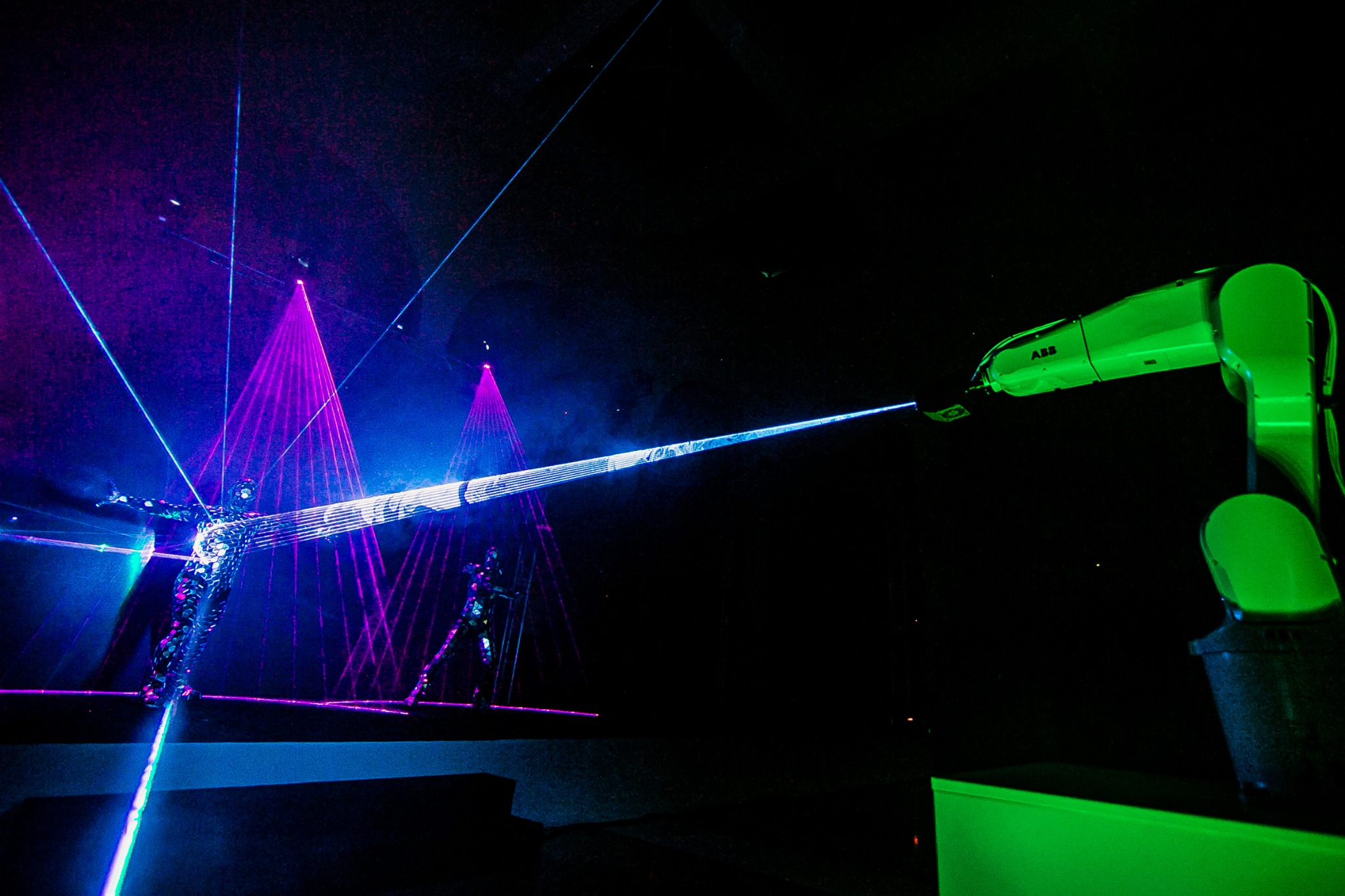 Interaktywny pokaz laserowy z udziałem robota przemysłowego ABB i tancerzy w kostiumach odbijających światło.