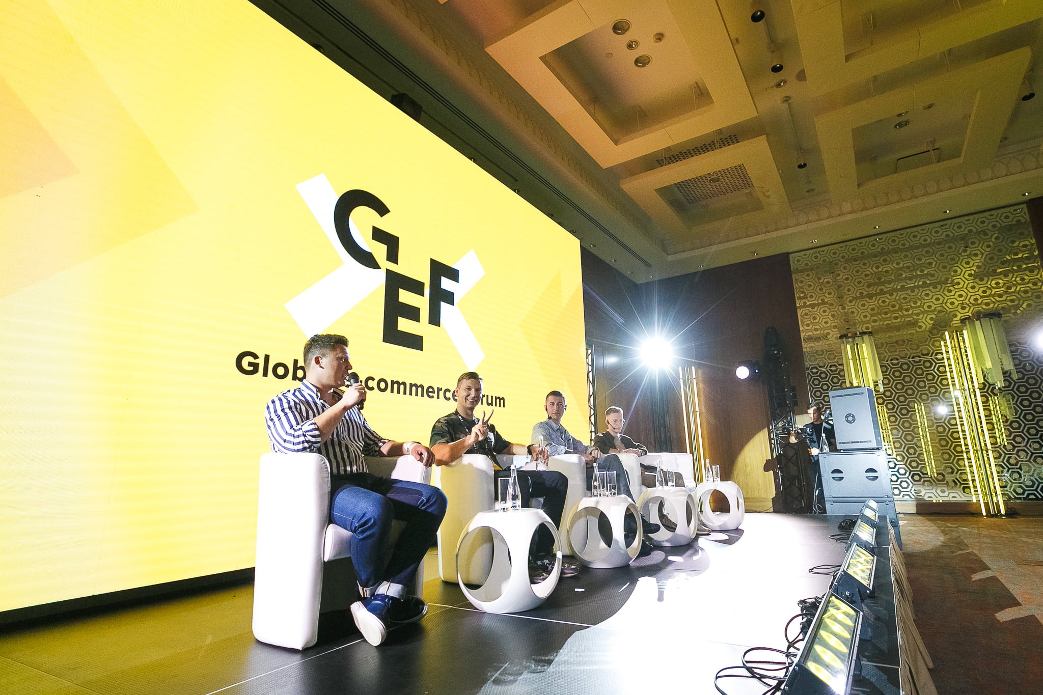 Panel dyskusyjny na konferencji Global Ecommerce Forum z czterema prelegentami siedzącymi na białych krzesłach, z logo GEF na żółtym tle, oświetlenie sceniczne i ozdobne ściany w tle.