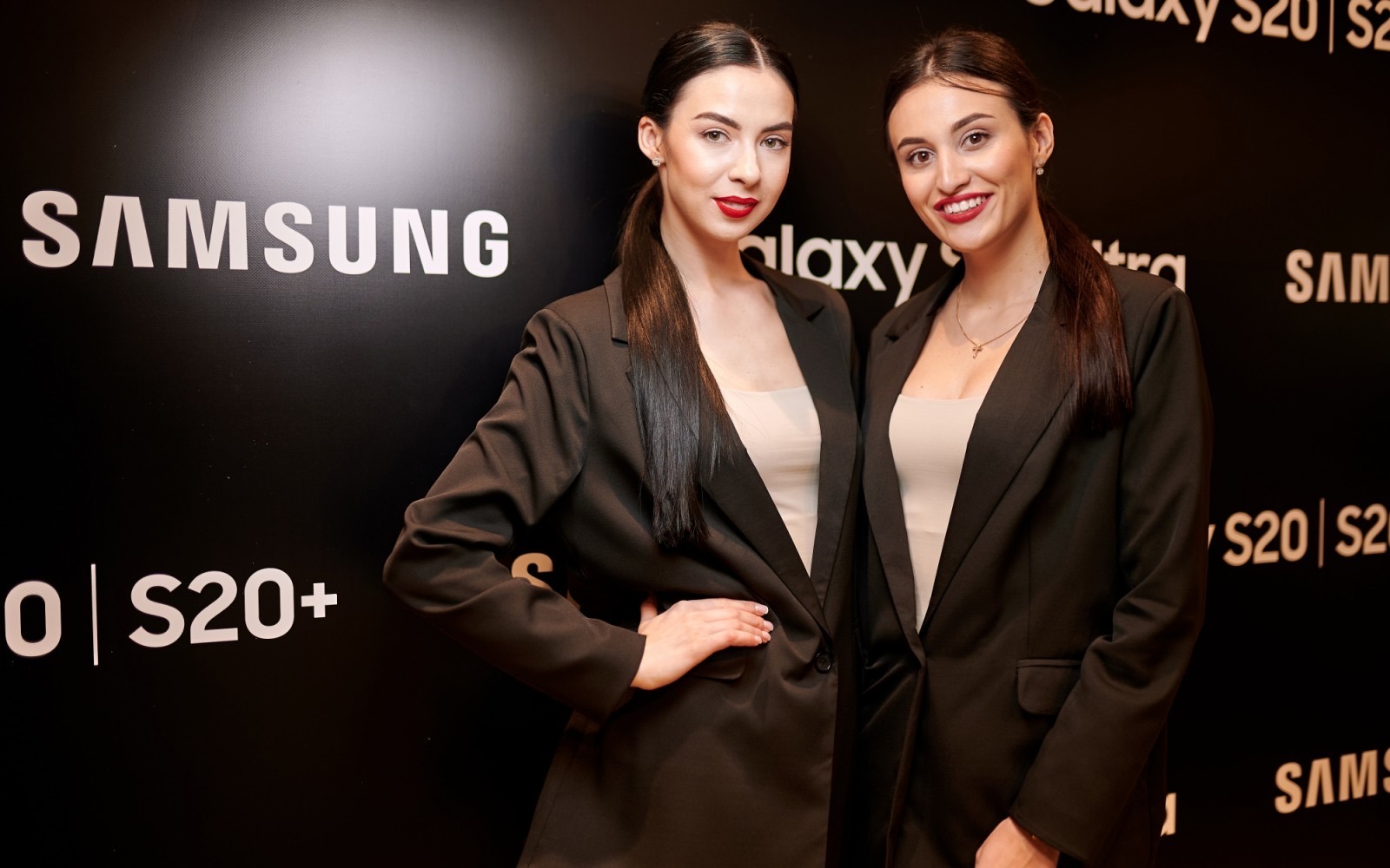 Dwie modelki w czarnych marynarkach pozują na tle z logo Samsung Galaxy S20 podczas wydarzenia promocyjnego.