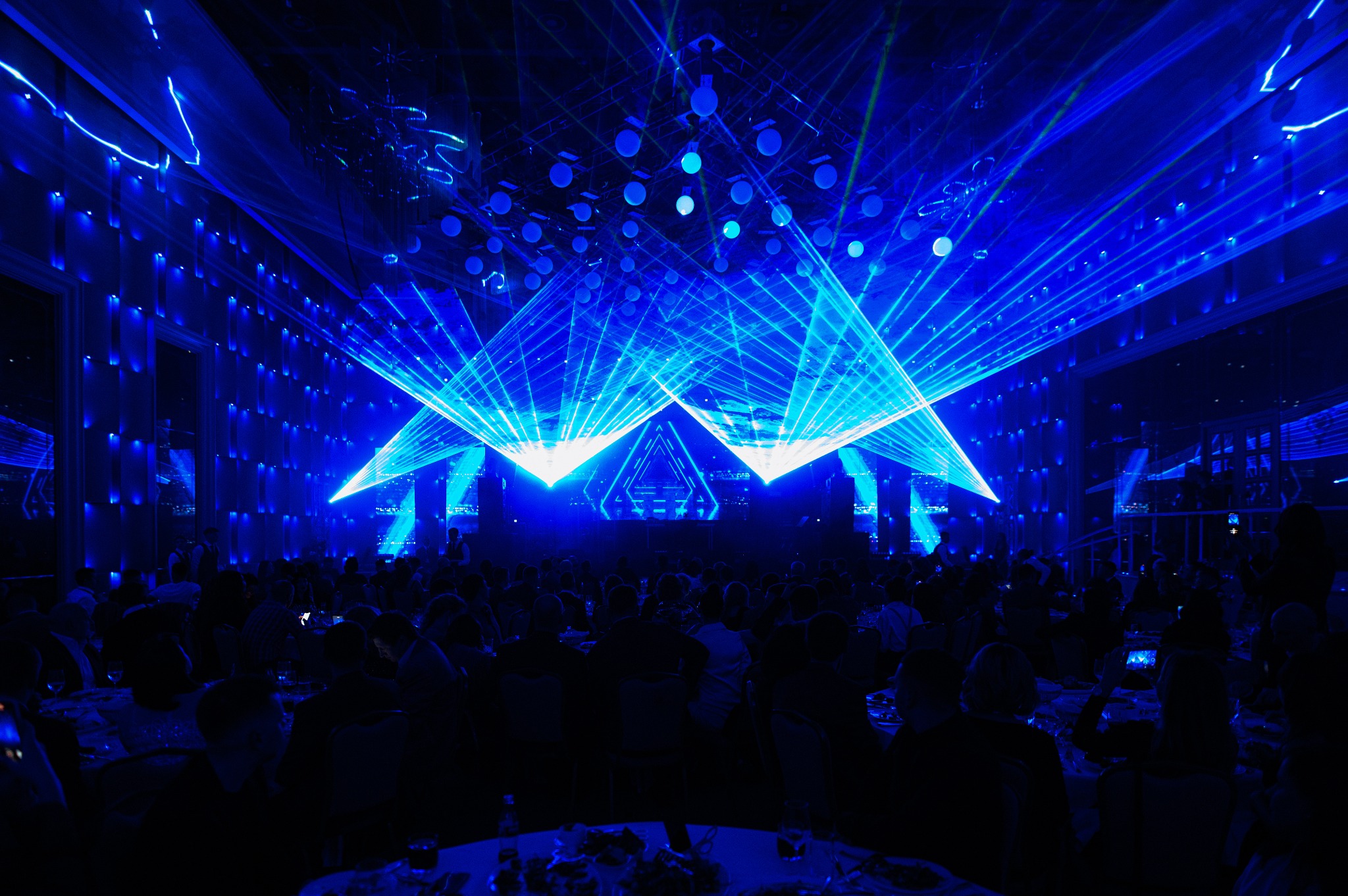 Sala bankietowa oświetlona intensywnym niebieskim światłem laserowym, z widocznymi sylwetkami gości siedzących przy stołach.