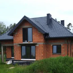 Surowy stan domu jednorodzinnego z ciemnoszarą dachówką karpiówką, czarnymi rynnami i oknami, widoczny mur z czerwonej cegły, na pierwszym planie zarośnięta trawa.