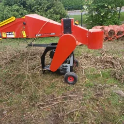 Czerwona rębarka do gałęzi RS-100 na trawie z porozrzucanymi i pociętymi gałęziami, w tle worki wypełnione zrębkami.