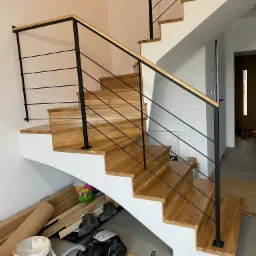 Drewniane schody z czarną, minimalistyczną balustradą z poziomych prętów, widok z dołu, z elementami wykończeniowymi w tle.