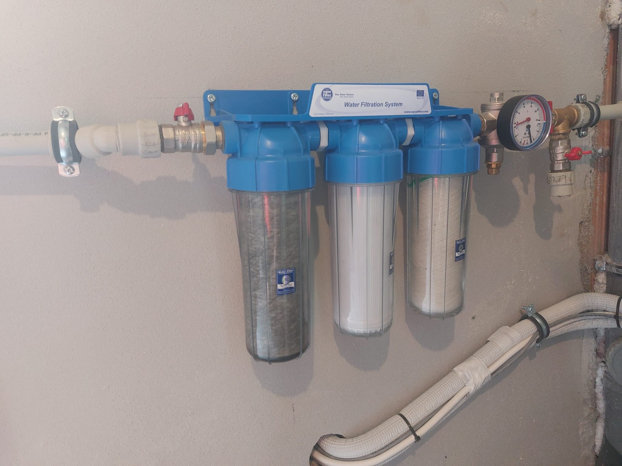 Trzystopniowy system filtracji wody zamontowany na ścianie, z widocznymi rurami doprowadzającymi i odprowadzającymi wodę oraz manometrem kontrolnym.