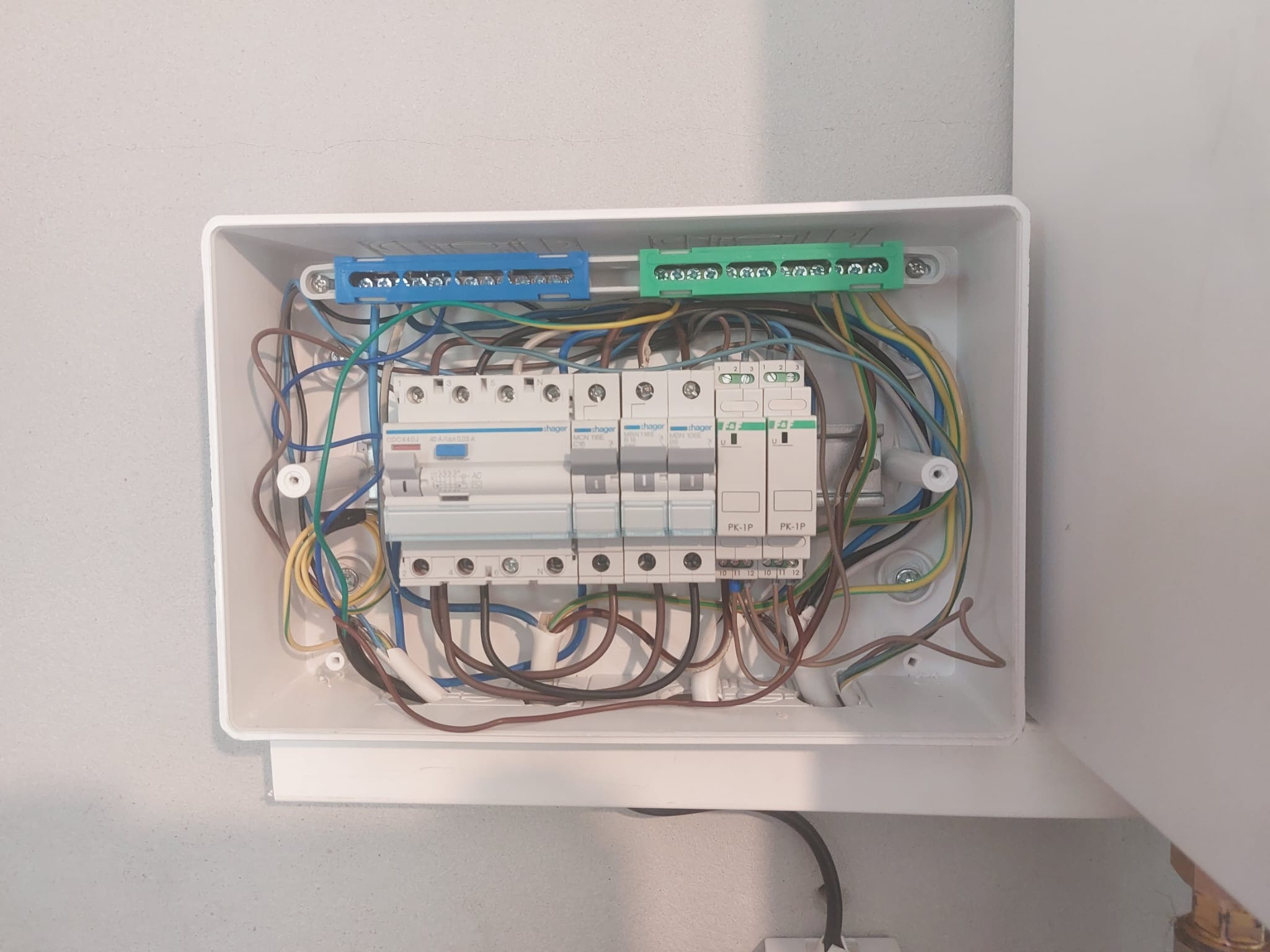 Otwarta skrzynka elektryczna z zainstalowanymi bezpiecznikami Hager i kolorowymi przewodami na tle szarej ściany.