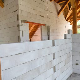 AD TOM BUD - Wznoszenie ściany z białych bloczków betonu komórkowego, widoczne spoiny i fragment drewnianej konstrukcji dachu w surowym stanie. Etap budowy domu.
