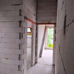 AD TOM BUD - Wnętrze w trakcie budowy: białe ściany z betonu komórkowego, widoczne otwory drzwiowe z nadprożami. W tle okno z widokiem na zieleń.