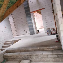 AD TOM BUD - Wnętrze w trakcie budowy: białe ściany z betonu komórkowego, drewniana konstrukcja dachu, widoczne worki z zaprawą, schody z bloczków betonowych, surowy stan wykończenia.