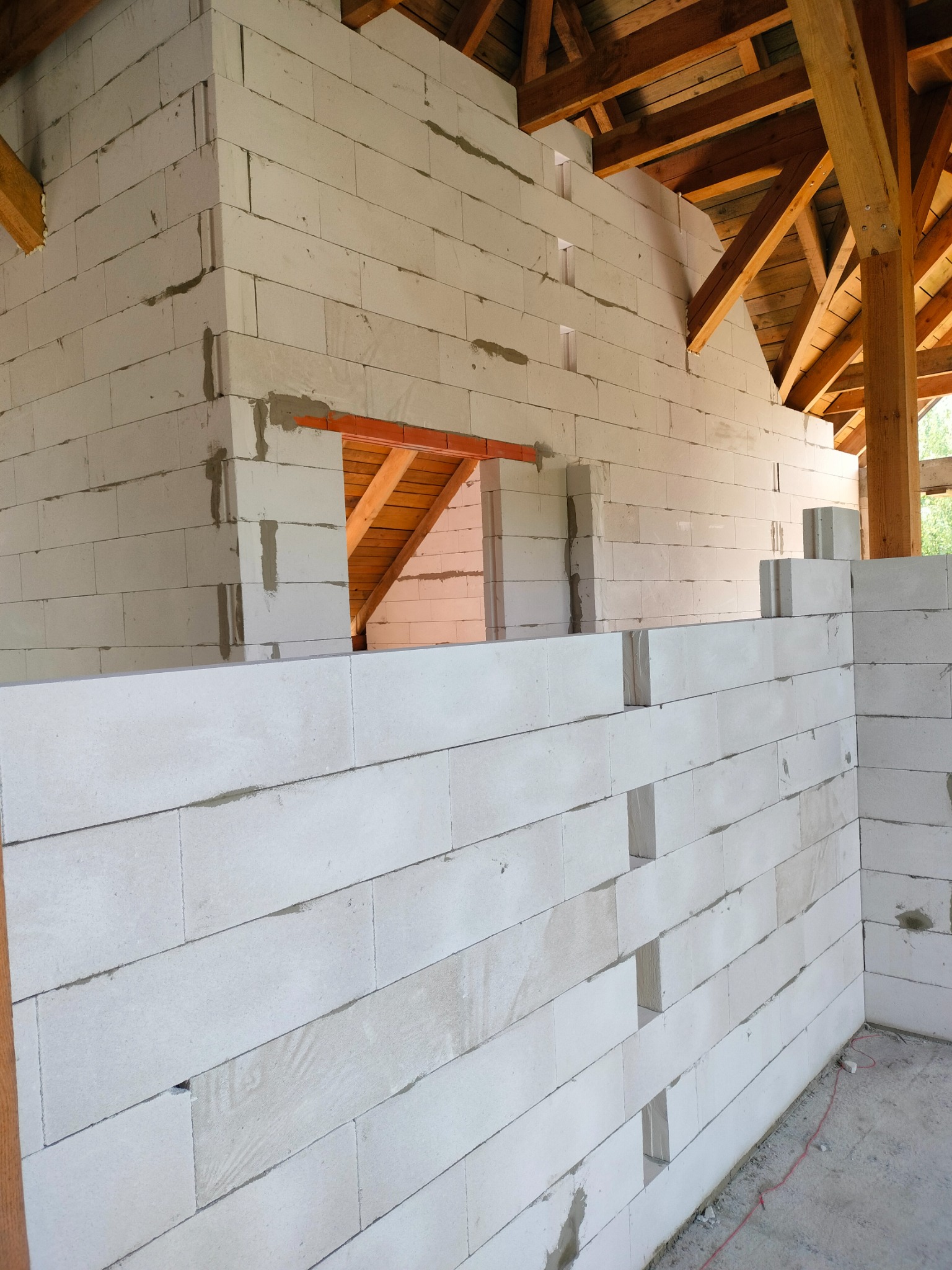Wznoszenie ściany z białych bloczków betonu komórkowego, widoczne spoiny i fragment drewnianej konstrukcji dachu w surowym stanie. Etap budowy domu.