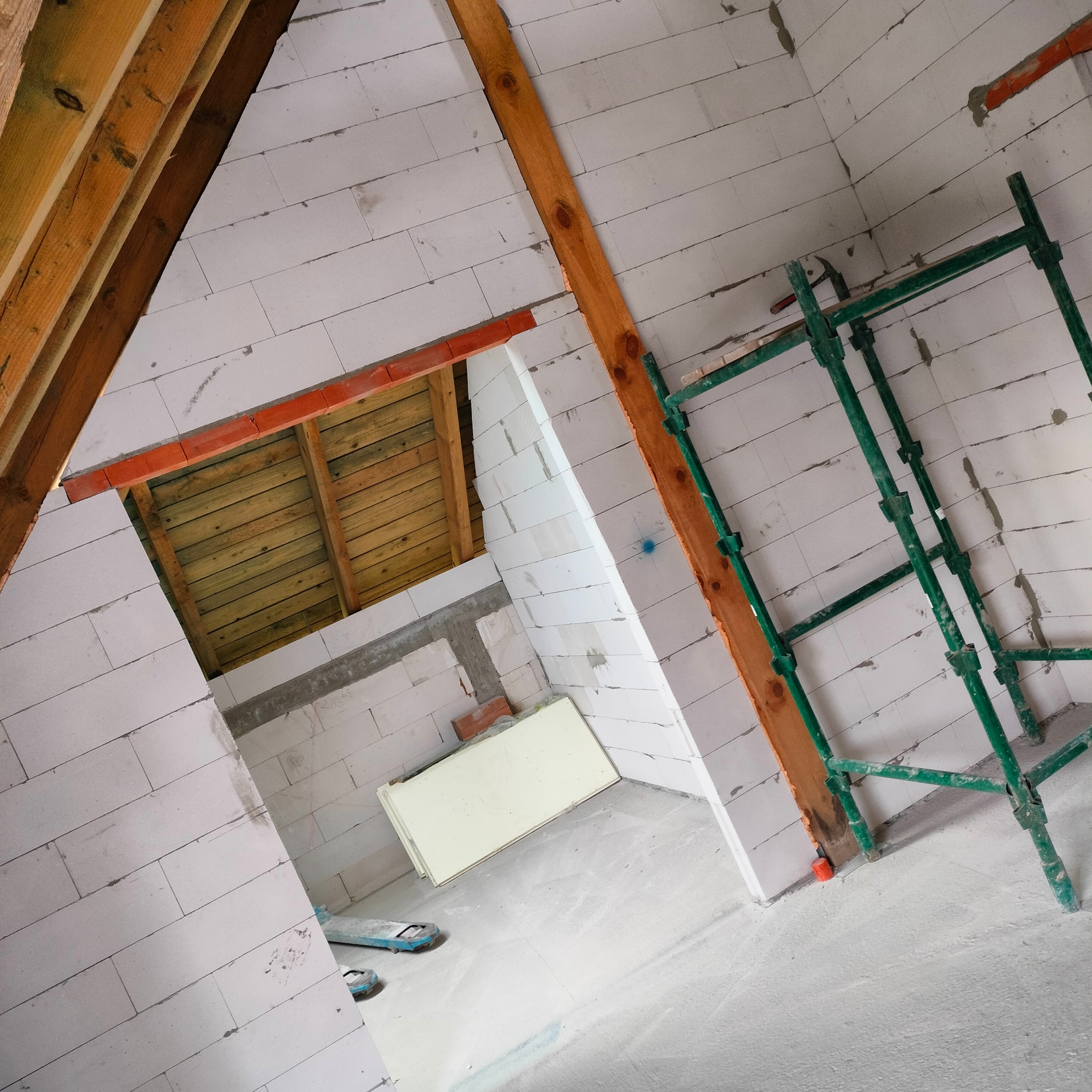 Wnętrze w trakcie murowania ścian z białych bloczków, widoczne drewniane elementy konstrukcyjne dachu i rusztowanie. Ukończony fragment ściany.