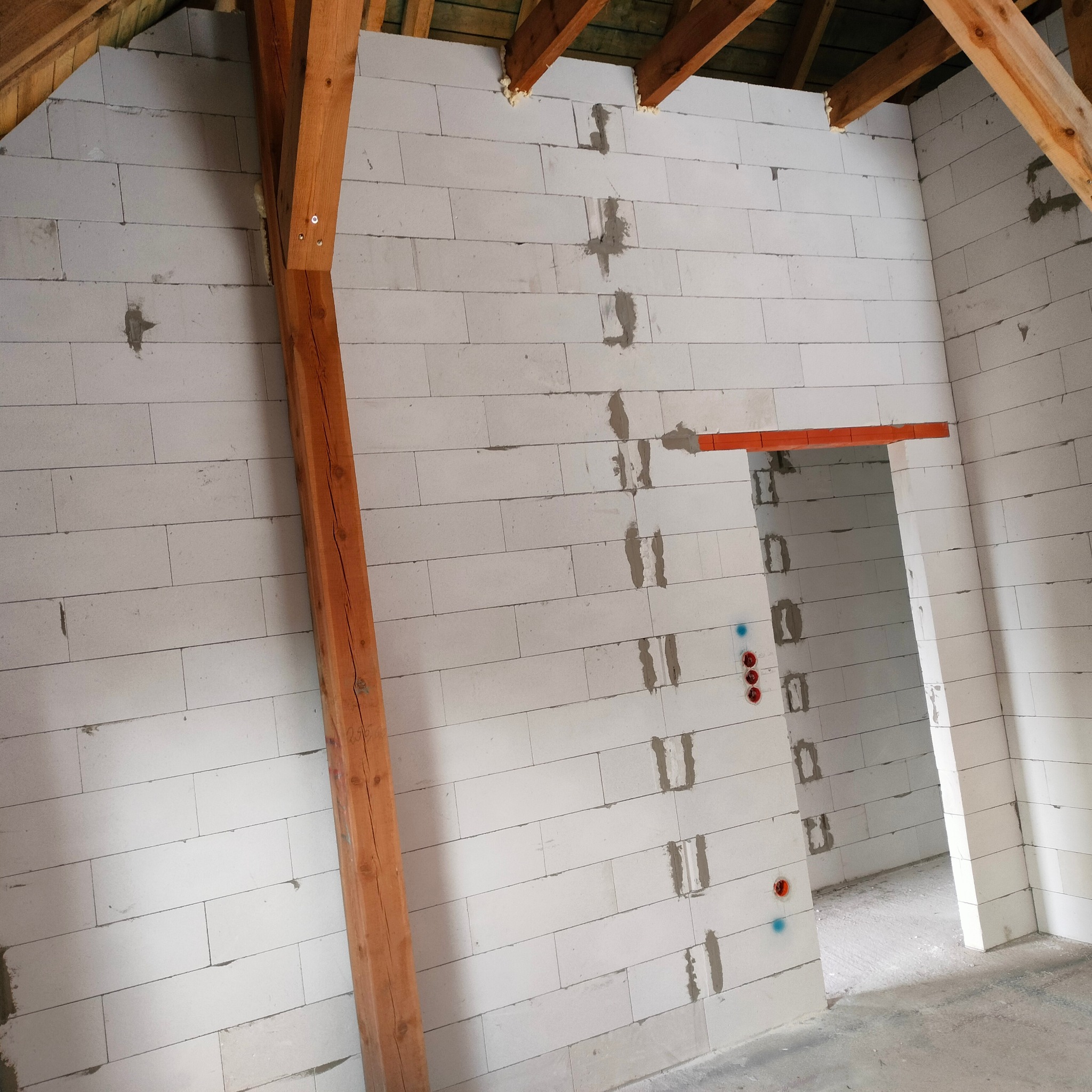 Wnętrze w trakcie budowy z białą ścianą z betonu komórkowego, drewnianą belką i widocznym otworem drzwiowym oraz instalacją elektryczną.
