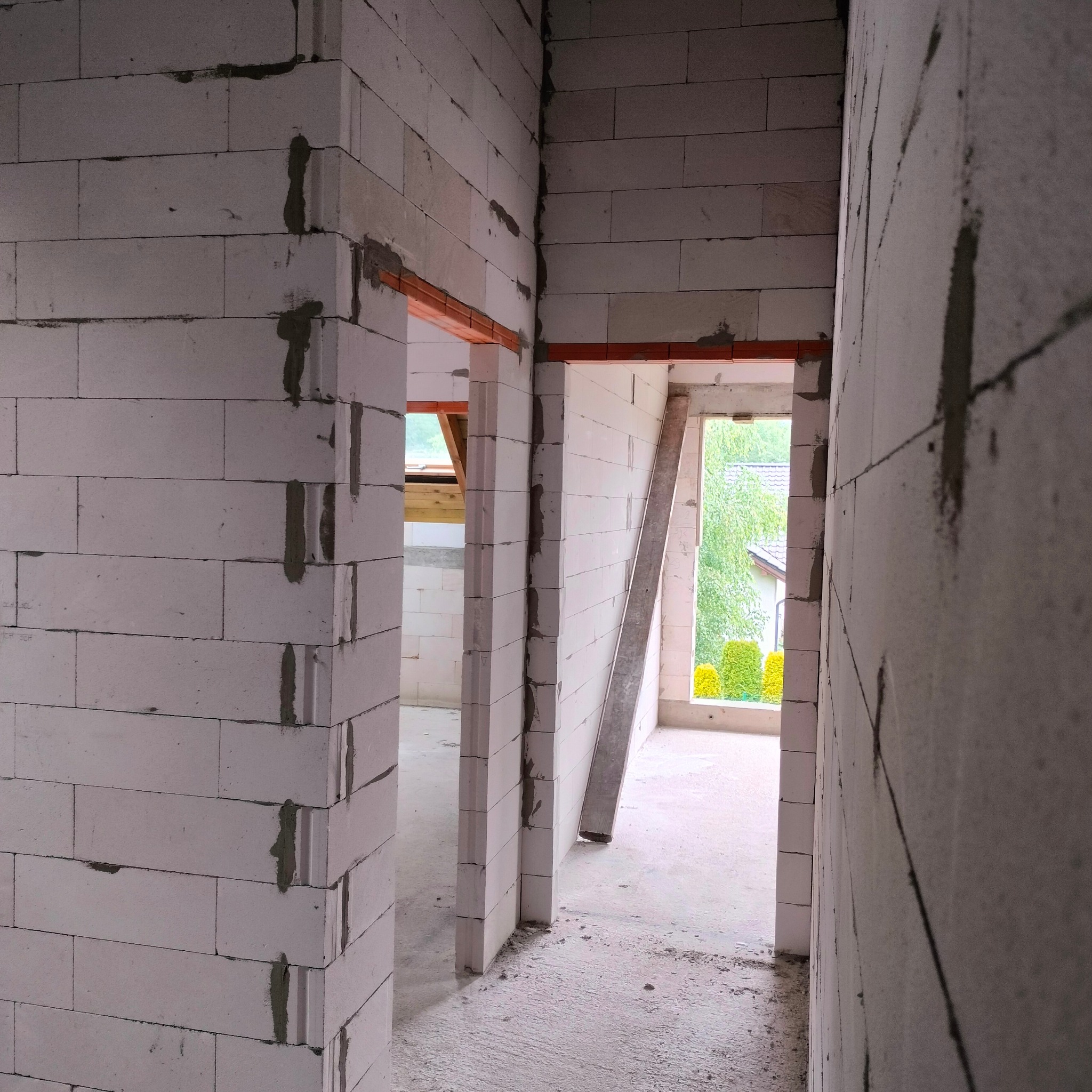 Wnętrze w trakcie budowy: białe ściany z betonu komórkowego, widoczne otwory drzwiowe z nadprożami. W tle okno z widokiem na zieleń.