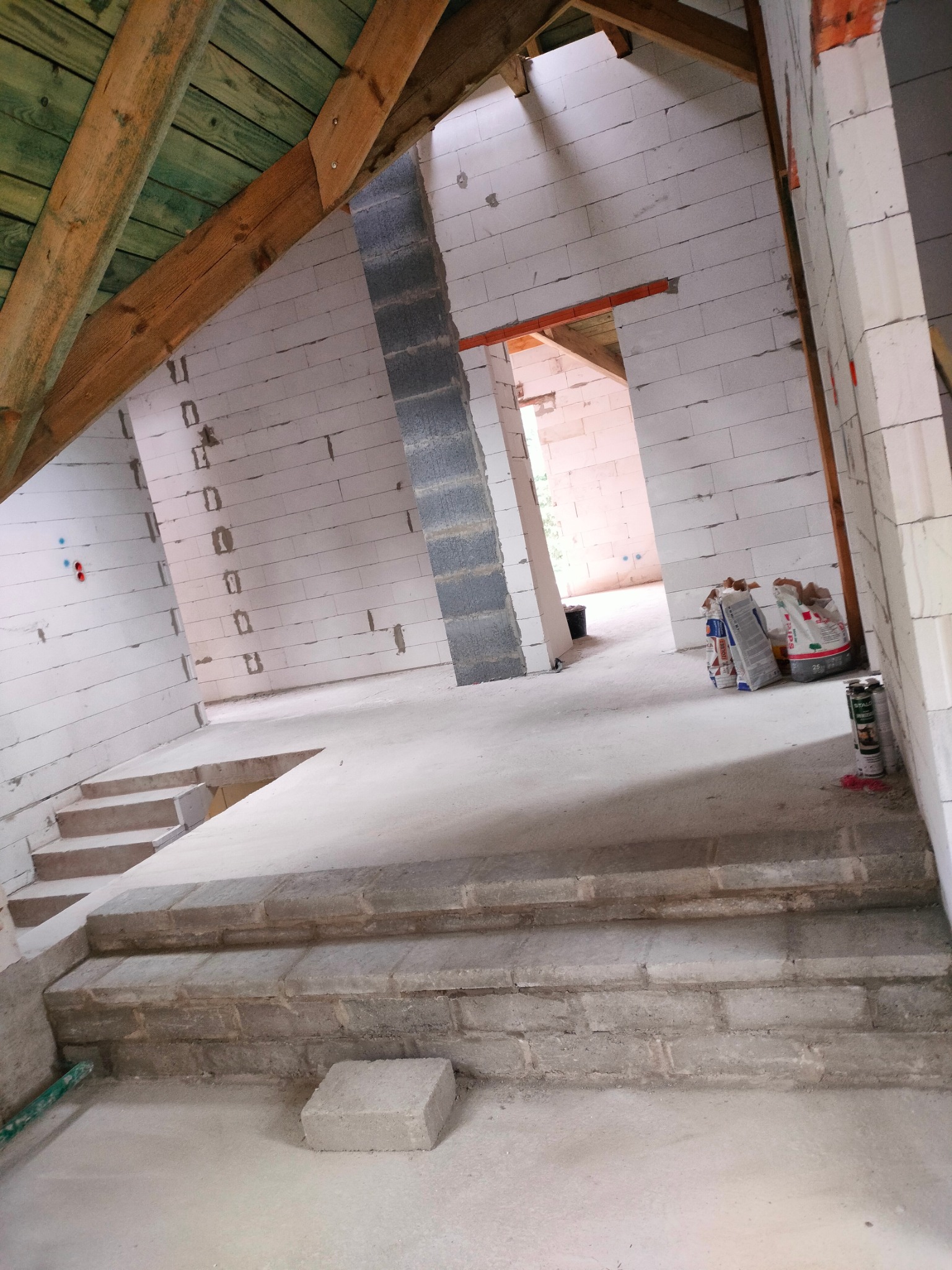 Wnętrze w trakcie budowy: białe ściany z betonu komórkowego, drewniana konstrukcja dachu, widoczne worki z zaprawą, schody z bloczków betonowych, surowy stan wykończenia.