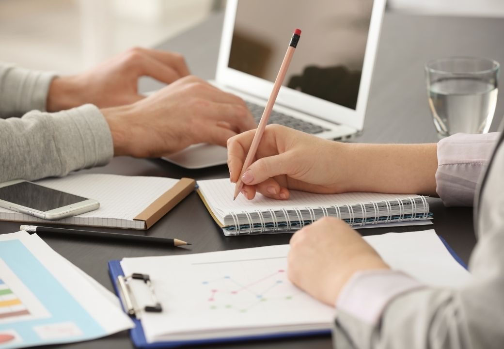Dwie osoby pracują przy biurku: jedna pisze ołówkiem w notatniku, druga używa laptopa. Widoczne są również smartfon, szklanka wody i dokumenty z wykresami.