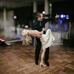 Grzegorz Żyła - Para tańczy na parkiecie podczas uroczystości. Mężczyzna w czarnym stroju trzyma kobietę w białej sukni w pozycji tanecznej, w tle widoczne światła i dekoracje.