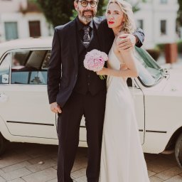 Grzegorz Żyła - Para młoda w eleganckich strojach pozuje przy kremowym, zabytkowym samochodzie z kwiatami. Pan młody w okularach, panna młoda z bukietem różowych piwonii.